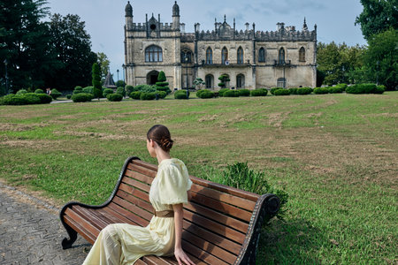 A woman in a pale yellow dress sits on a wooden bench in a manicured park, facing a grand castle like building, evoking quiet elegance and scenic charm.の写真素材