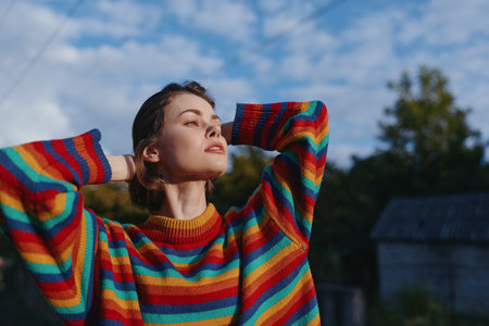 Young adult woman in a colorful rainbow sweater outdoors, relaxed and looking up with hands behind head. Serene expression and sunlight portrait for lifestyle, fashion and travel use.の写真素材