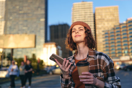 Woman holds coffee and phone in a city street at golden hour, candid authenticity and mindful living; emotional storytelling portrait with warm glow, urban lifestyle and relaxed confidence.の写真素材