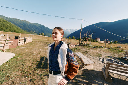 Woman in mountains village standing outdoors with backpack and jacket, sunlight on face and eyes closed enjoying nature and travel, casual style in peaceful rural landscape.の写真素材