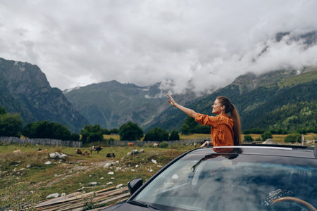 A woman leans from a sunroof of a car in a rural valley, pointing toward distant mountains, capturing a moment of adventure, freedom, and exploration in open countrysideの写真素材