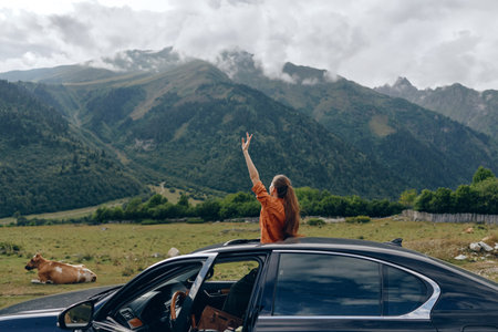 woman car mountains meadow cow roadtrip nature travel sunroof, woman standing through sunroof in meadow with cow and peaks, authentic inclusivity joyful freedom relaxed smileの写真素材