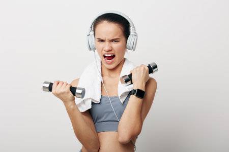 Fitness woman lifting dumbbells with intensity, wearing sports bra and headphones, towel over shoulder in a clean studio, focused energy and powerful workout moment.の写真素材