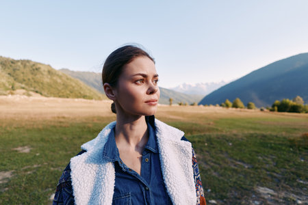 Woman portrait in nature with mountains and meadow, outdoors travel scene. Thoughtful young female stands in wide landscape at golden hour wearing denim and fleece jacket for adventure.の写真素材