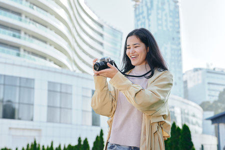 Candid shot of a smiling woman photographer outdoors in an urban setting, camera in hand, light jacket, modern buildings in background, travel and creative photography vibe.の写真素材