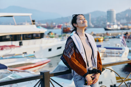 Woman enjoying seaside breeze in a textured coat and scarf by a marina, bright sunlight, distant hills, and boats, creating a stylish travel mood and relaxed lifestyle sceneの写真素材