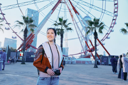 Ferris wheel amusement park scene with a smiling woman holding a camera, outdoor fashion, palm trees in the background, evening lights, candid travel photography and explorationの写真素材