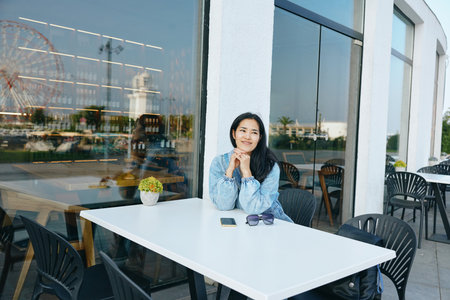 Woman sits at a white outdoor cafe table with thoughtful expression, glass facade reflections, sunny day, small plant, sunglasses on table, casual denim shirt, relaxed urban cafe vibeの写真素材