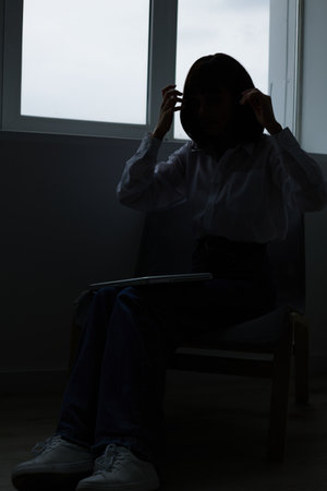 A solitary silhouette sits by a window in dim light, creating a moody scene that conveys reflection, silence, and gentle anxiety, inviting curiosity about a hidden story.の写真素材