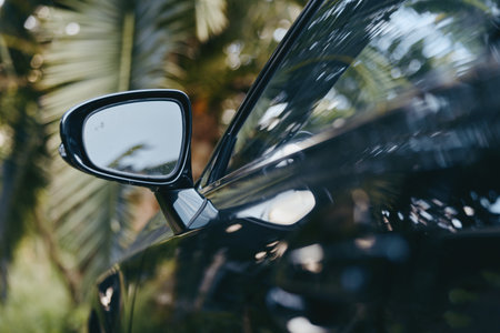 Car side mirror reflection with palm tree on glossy black car window, closeup shot of modern vehicle exterior showing shiny paint, mirror detail and outdoor tropical moodの写真素材