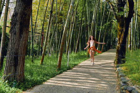 Woman running on a gravel path through a dense bamboo forest, wearing a flowing dress with arms outstretched, joyful expression and sunlight filtering through tall trunks and green foliage.の写真素材