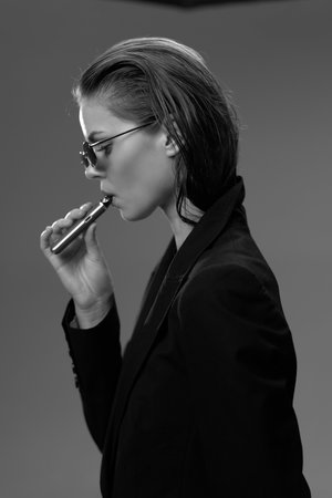 Black and white portrait of a woman vaping with sunglasses and slicked-back hair in a dark blazer.