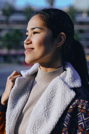 Woman in a shearling coat outdoors, bathed in warm sunlight, gazing to the side with a calm smile,...