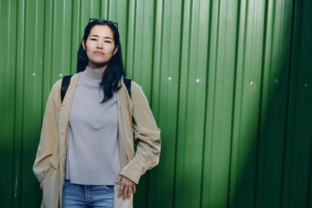 Woman standing before a green metal wall in a beige coat and gray knit sweater, backpack ready for urban exploration, casual fashion and street style shot highlighting everyday attire.の写真素材