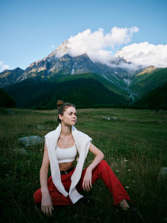 Outdoor scene with a woman seated in a grassy meadow, mountains above and clouds in a clear blue...