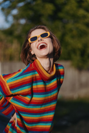 Woman laughing young adult in a colorful striped sweater and orange sunglasses, casual outfit outdoors. Joyful portrait with bob haircut, playful pose and vibrant lifestyle fashion vibe in sunlight.の写真素材