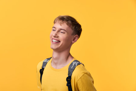 Young man wearing a yellow shirt and backpack, smiling against a bright yellow background, conveying optimism, energy, and casual everyday styleの写真素材