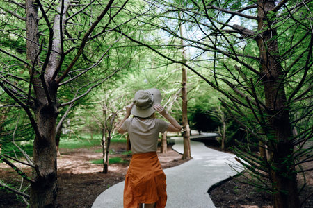 Woman hat path forest trees nature walking trail captured from back view on a winding route, casual summer outfit with orange skirt, peaceful outdoor exploration amid lush greenery.の写真素材