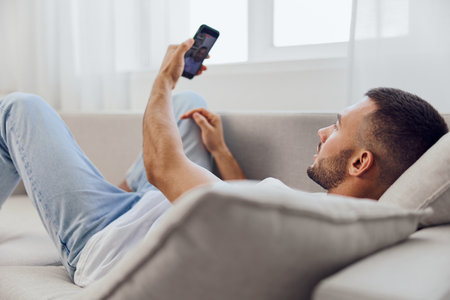Relaxed man enjoying a video call at home, wearing casual attire, showcasing comfort and modern lifestyle in a bright living room setting.の写真素材