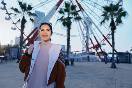 Woman traveler outdoors near a ferris wheel, smiling in a warm coat, enjoying urban leisure, palms sway in the breeze, evening light enhances candid street scene, modern entertainment backdrop forの写真素材