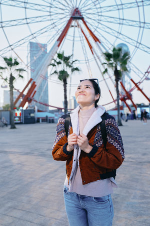 Ferris wheel in the background as a woman stands outdoors wearing a cozy jacket and sweater, looking...