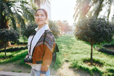 Photography of a woman outdoors in a park with a camera, wearing a patterned jacket and backpack, enjoying a sunny day. Casual fashion and travel mood in bright light.の写真素材