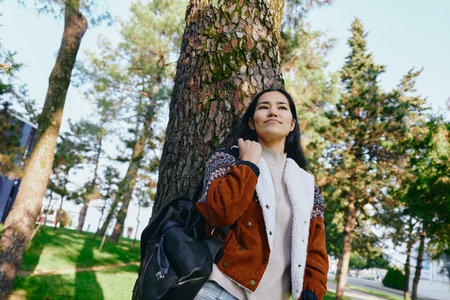 woman outdoors tree park serenity daytime casual nature with backpack jacket sunlit scene for lifestyle travel wellness and nature themesの写真素材