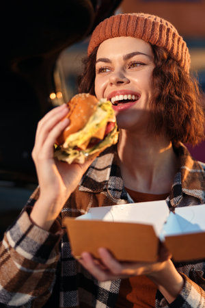 Woman smiling as she enjoys a burger and street food outdoors, urban evening takeout moment with beanie and plaid jacket. Candid lifestyle shot with authenticity, golden hour glow and emotionalの写真素材