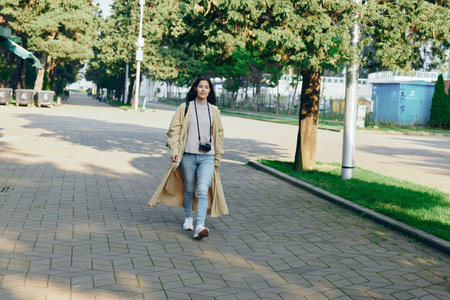 A stylish woman walks along a park path wearing a long coat and carrying a camera, exploring...