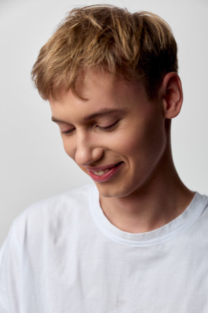 Boy with a gentle smile in a white t-shirt looking down, conveying quiet happiness in a casual mood,...
