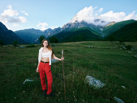 Woman with a wooden staff stands in a green meadow beneath towering mountains, wearing a white...