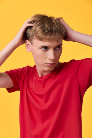 Boy with hands on head in a red shirt against a bright yellow backdrop, conveying confusion and...