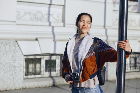 Camera in hand, smiling woman stands outdoors beside a pole, urban backdrop, casual fashion vibe for...