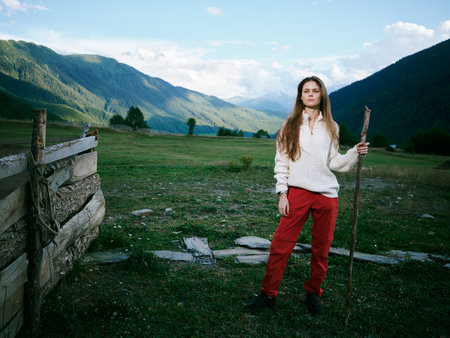 Woman stands in a green valley with distant mountains, holding a wooden staff, wearing a white...