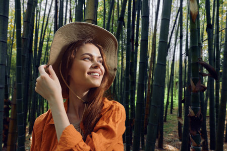 Woman smile hat bamboo forest portrait of a young traveler in nature, wearing straw hat and orange shirt, looking joyful among tall green stalks, outdoor lifestyle and eco travel.の写真素材