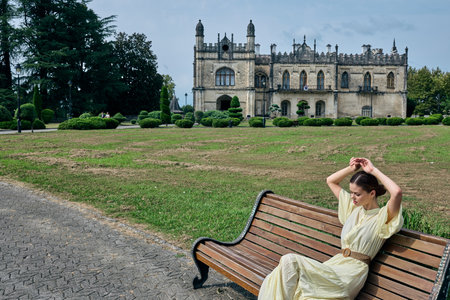 Woman in a light dress sits on a wooden bench in a park with a grand historic castle in the background, creating a serene travel and sightseeing moment.の写真素材