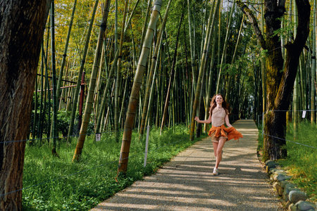 Woman girl bamboo forest path running in nature wearing an orange skirt, joyful motion and sunlight filtering through tall bamboo trees on a gravel trail for outdoor lifestyle.の写真素材