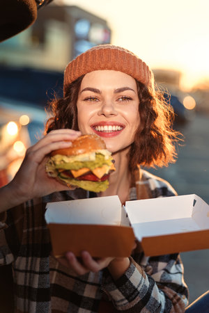 Woman with beanie and plaid jacket smiling while holding burger outdoors at golden hour, candid lifestyle portrait evoking authenticity, mindful living and emotional storytelling in urban sunset.の写真素材