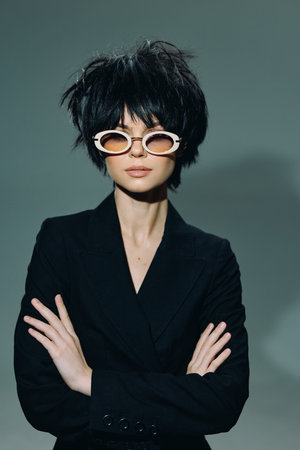 Stylish woman wearing round sunglasses and black blazer with crossed arms against plain background.の写真素材