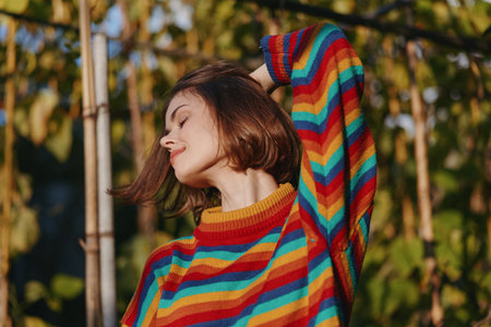 Woman portrait in colorful striped sweater smiling with eyes closed outdoors in warm sunlight, short brown hair, peaceful expression and autumn foliage background lifestyle.の写真素材
