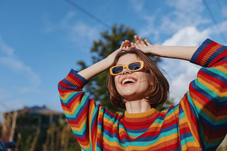 Woman smile sunglasses sweater happy portrait outdoors sunshine in colorful striped jumper, cheerful young adult enjoying a sunny day with hands raised and looking up at sky.の写真素材