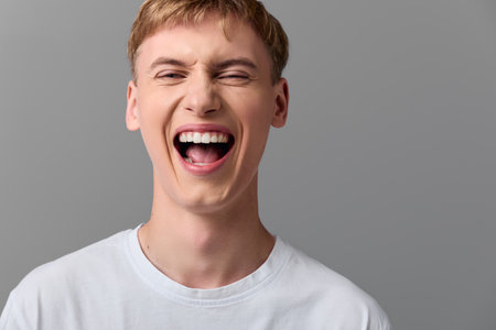 Laughter, open mouth smile, energetic young man wearing white tshirt in studio, conveying joy and...