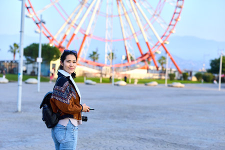 Woman with camera at an outdoor park, standing near a towering ferris wheel, wearing a casual jacket...