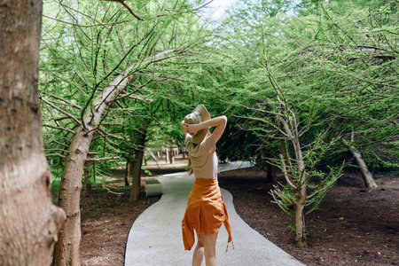Woman walking on gravel path in forest wearing straw hat and orange skirt, back view. Leisurely summer stroll among green trees, natural light and peaceful nature scene outdoors.の写真素材