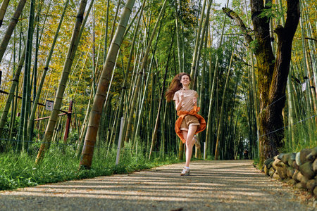 Woman running on a forest path through tall bamboo, smiling in motion wearing orange skirt and casual top. Outdoor nature scene with sunlight, trees and peaceful trail.の写真素材