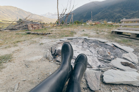 boots and firepit with cold ashes in foreground, mountains and rural campsite in background, camping outdoors with wooden bench, person relaxing in rubber bootsの写真素材