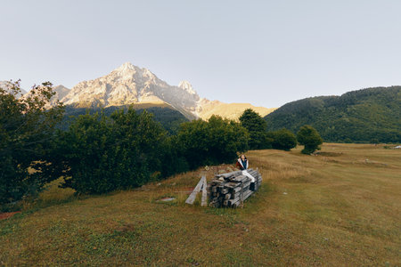 Mountain meadow with hiker and backpack sitting on a stonewall, trees and distant peaks at sunrise....