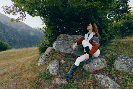 Woman sitting on rocks under a tree in a mountain meadow with mountains in the background, nature...
