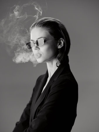 Black and white portrait of a stylish woman wearing sunglasses exhaling smoke in a minimalistic studio setting.の写真素材