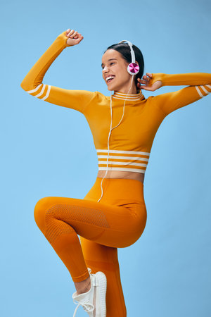 Woman in orange crop top and leggings performing energetic aerobic pose with raised knee and fists,...